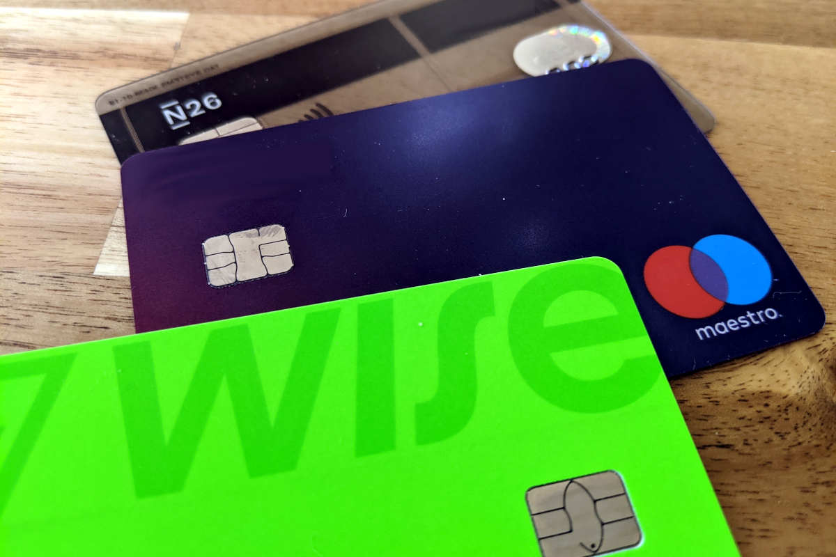 3 payment cards on a wooden background: a green Wise card, a blue Maestro card and a transparent N26 card