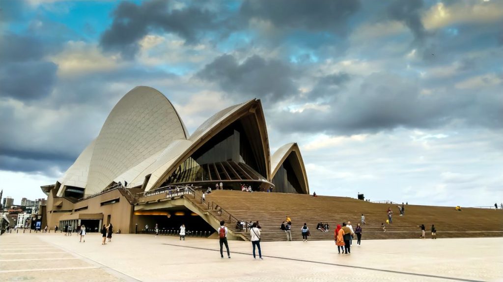 Sydney Opera House - people walking around and sitting on the steps. Clouds above.