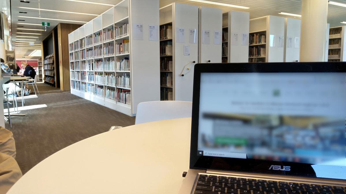 View of laptop on table in library for remote work. Book cases in the distance