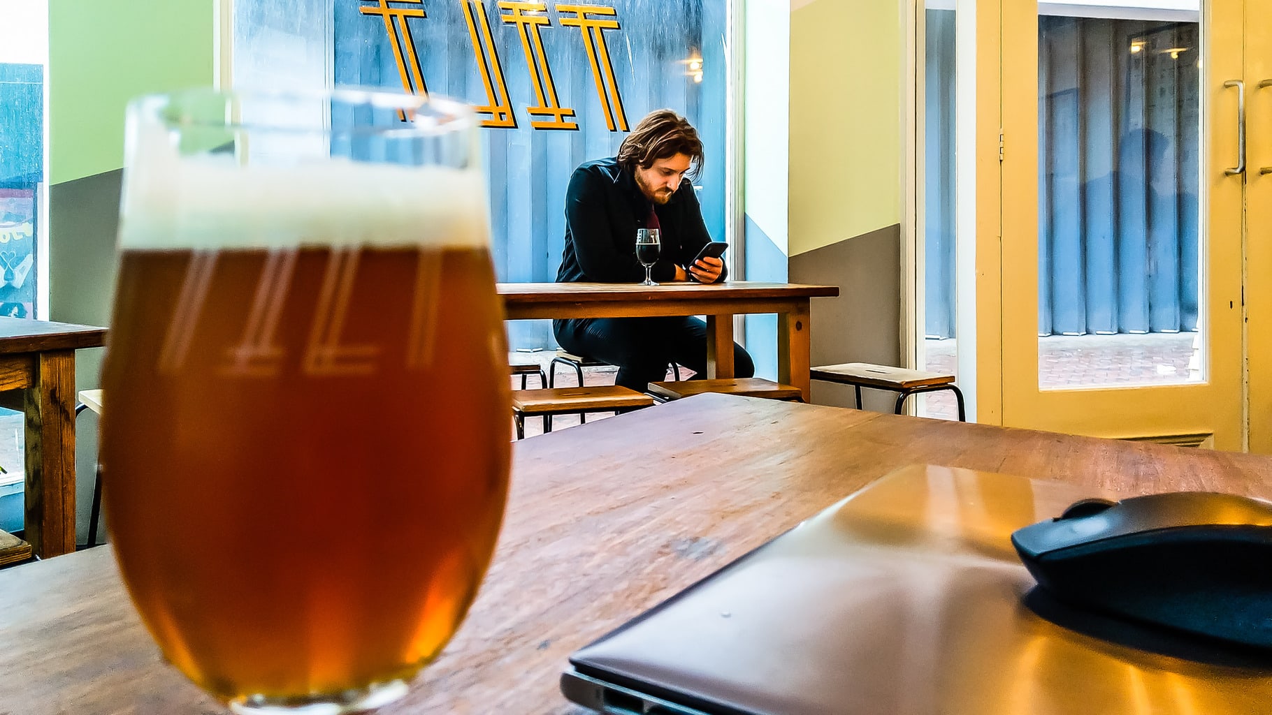 Close up of craft beer in glass and laptop with person on smartphone in the background